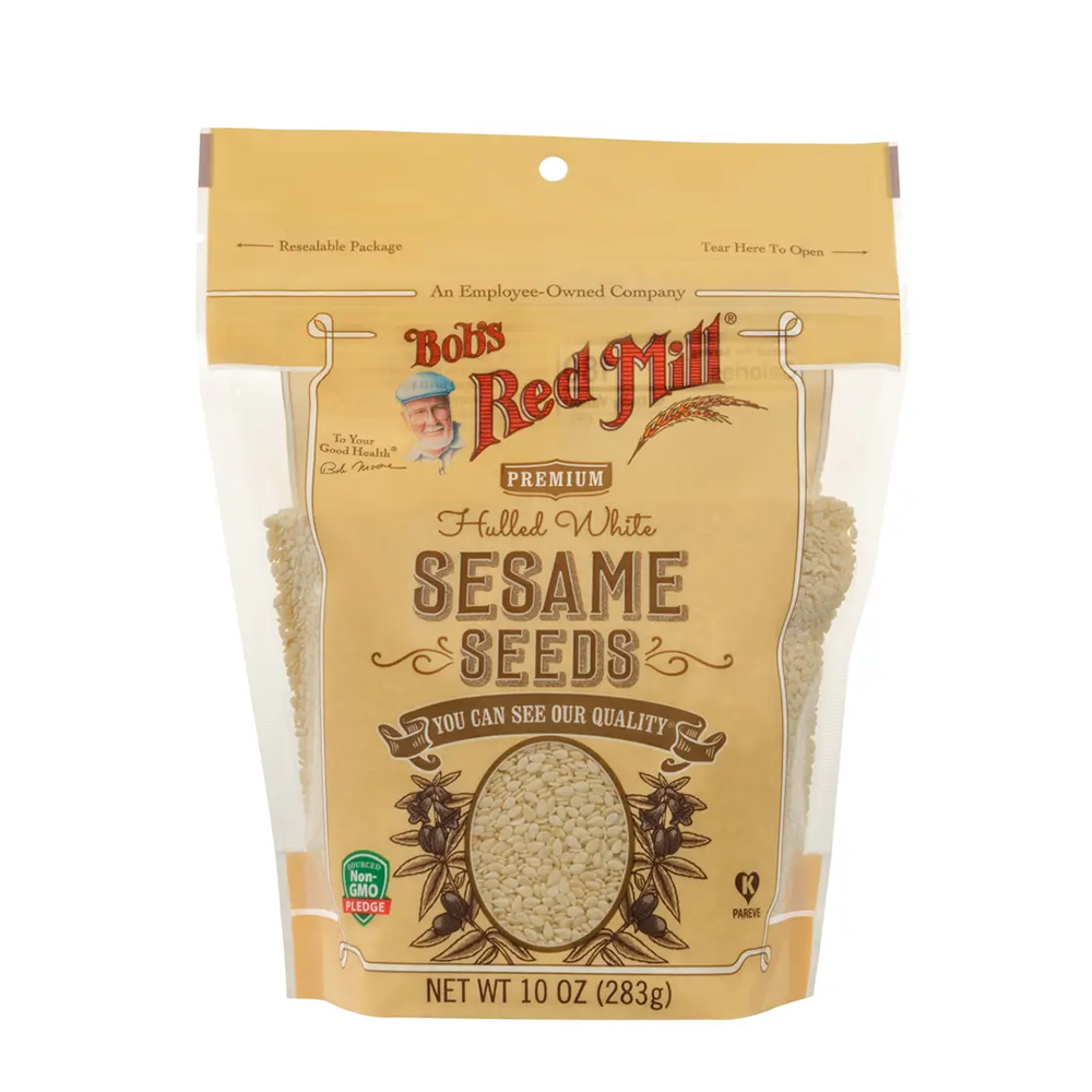 A 10 oz bag of Bob's Red Mill Premium Halled White Sesame Seeds, featuring a tan stand-up pouch with clear windows showing the light beige seeds inside.