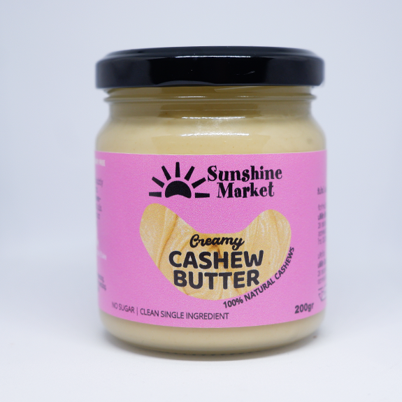 A jar of Sunshine Market Cashew Butter with a pink label, displaying the product name and the claim of being creamy, with a cashew image, indicating it is made from 100% natural cashews.