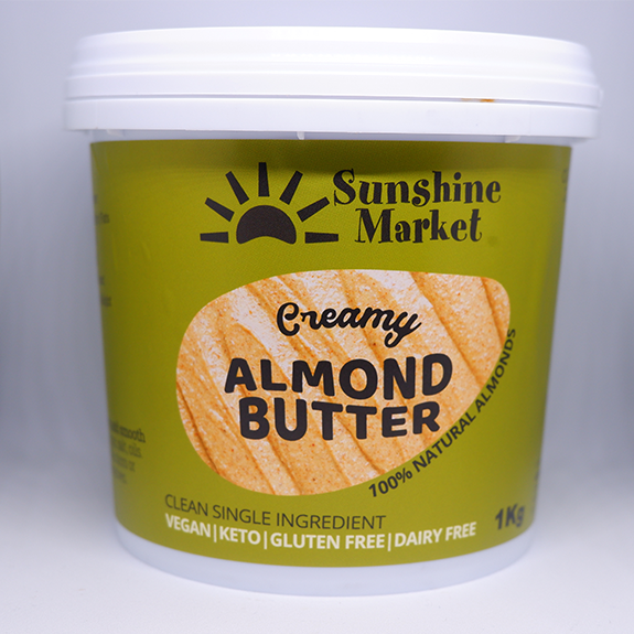 A container of Sunshine Market Almond Butter with the label showing the brand name and the product name, creamy almond butter, and the weight of 1kg. The container is green with a picture of a slice of bread with almond butter on it.