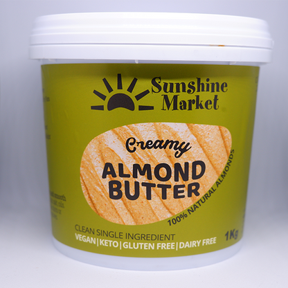 A container of Sunshine Market Almond Butter with the label showing the brand name and the product name, creamy almond butter, and the weight of 1kg. The container is green with a picture of a slice of bread with almond butter on it.