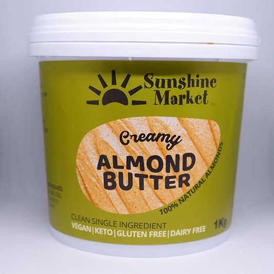 A container of Sunshine Market Almond Butter with the label showing the brand name and the product name, creamy almond butter, and the weight of 1kg. The container is green with a picture of a slice of bread with almond butter on it.